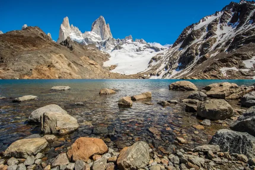 Crystal-clear mountain lake with rocky shores and snow-covered granite peaks in Patagonia, set against a bright blue sky.