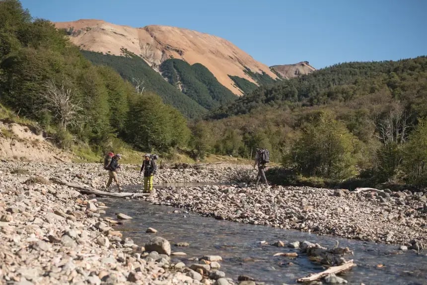 Hikers crossing a shallow river in a rocky valley with forested hills and striking orange mountains in the background