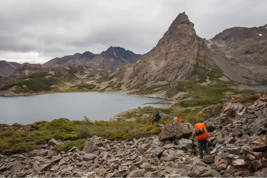 Hikers with backpacks climbing over rocky terrain toward a mountain lake in the remote Dientes de Navarino range on a cloudy day